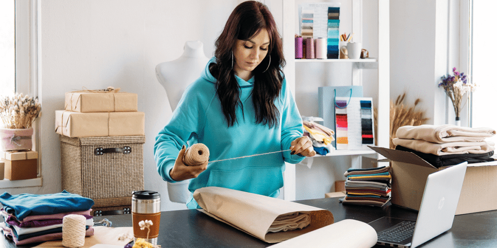 How to Make Money on Etsy in 2026: A Complete Beginner's Guide features an aspiring online entrepreneur managing a successful Etsy shop from a modern home office. A laptop displays an Etsy storefront with handmade crafts, digital downloads, personalized gifts, and vintage products. The workspace includes packaging materials, creative tools, a notebook, and a smartphone showing new order notifications. Sales graphs, customer reviews, and shipping labels illustrate business growth and daily operations. The background highlights a productive and organized environment that reflects the flexibility of running an online business from anywhere. The image represents creativity, entrepreneurship, and financial independence while showcasing the opportunities Etsy offers beginners. It emphasizes building a profitable online store through quality products, effective marketing, excellent customer service, and consistent effort in 2026.