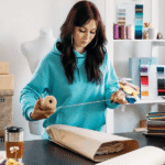 How to Make Money on Etsy in 2026: A Complete Beginner's Guide features an aspiring online entrepreneur managing a successful Etsy shop from a modern home office. A laptop displays an Etsy storefront with handmade crafts, digital downloads, personalized gifts, and vintage products. The workspace includes packaging materials, creative tools, a notebook, and a smartphone showing new order notifications. Sales graphs, customer reviews, and shipping labels illustrate business growth and daily operations. The background highlights a productive and organized environment that reflects the flexibility of running an online business from anywhere. The image represents creativity, entrepreneurship, and financial independence while showcasing the opportunities Etsy offers beginners. It emphasizes building a profitable online store through quality products, effective marketing, excellent customer service, and consistent effort in 2026.
