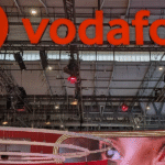 A modern smartphone connected to the Vodafone UK 5G network, displaying ultra-fast mobile connectivity in a smart city environment. The image features a professional using a 5G-enabled device while working remotely, streaming high-definition content, and accessing cloud-based applications. Digital graphics illustrate wireless signals, network coverage, smart technology, and secure data transmission. The vibrant urban background highlights connected buildings, autonomous technologies, and seamless communication powered by Vodafone UK's advanced 5G infrastructure. The scene reflects the future of mobile innovation, demonstrating how Vodafone UK's 5G technology delivers faster speeds, lower latency, reliable connectivity, and enhanced digital experiences. This visual represents modern communication, intelligent networking, and the evolution of mobile technology that supports everyday life, business productivity, entertainment, and smart digital living across the United Kingdom.
